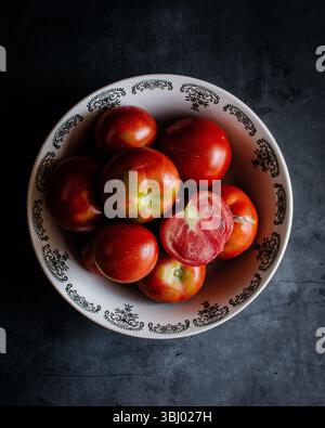 Ein Blick von oben nach unten auf frische, lebendige Tomaten in einer dekorativen Keramikschale vor dunklem Hintergrund. Eine Tomate wird in Scheiben geschnitten und zeigt ihre saftige Note Stockfoto