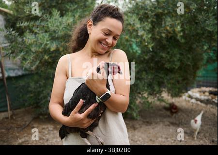 Lächelnde, glückliche Farmerin, die Freilandhennen hält und streichelt, in einer Scheune steht und Hühner und Hähne in der Öko-Farm füttert. Ökologischer Landbau Stockfoto
