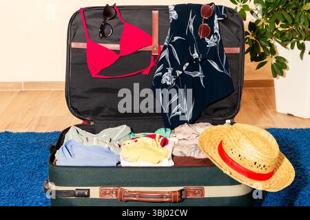 Offener Koffer mit Sommerkleidung, Sonnenbrille und Strohhut auf blauem Teppich, bereit für den Urlaub. Der Koffer ist für eine sonnige Sonne gepackt Stockfoto