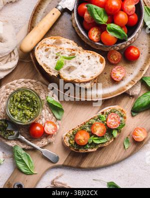 Rustikale Toasts mit lebhaften Kirschtomaten und duftendem Pesto, präsentiert auf einem Holzbrett. Frische Basilikumblätter und ein Messer ergänzen die Mediterra Stockfoto