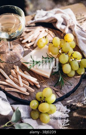 Ein ruhiges Stillleben im Freien mit einem Glas Wein, grünen Trauben, Rosmarin und Käse auf einem rustikalen Holzbrett. Sonnenlicht hebt die Strukturen und hervor Stockfoto