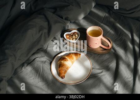 Ein goldenes hausgemachtes Croissant liegt auf einem weißen Teller, begleitet von einem Holztablett mit einer rosafarbenen Tasse Tee und einer herzförmigen Schüssel mit Nüssen, alles auf Stockfoto