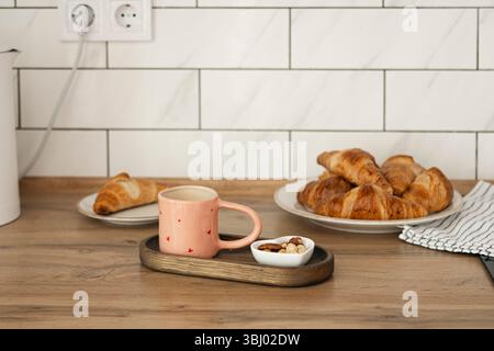 Frisch gebackene hausgemachte Croissants auf einem Holztisch mit einem gemütlichen rosa Becher und einer kleinen Schüssel Nüsse. Ein perfekter Start in einen entspannten Morgen in Hom Stockfoto