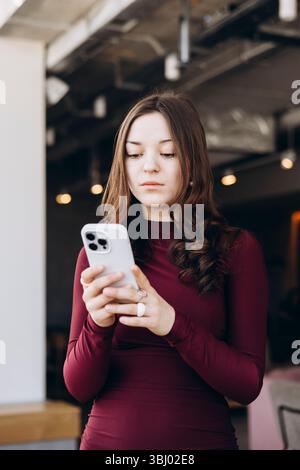 Eine junge Studentin mit langen Haaren lächelt, während sie in einem kastanienbraunen Kleid auf ihr Smartphone schaut. Sie ist drinnen und scheint sich zu amüsieren Stockfoto