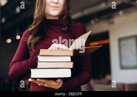 Eine Schülerin, die einen Stapel Bücher hält, in einem kastanienbraunen Oberteil gekleidet, durch ein Buch blättert und in einem warmen, gut beleuchteten Li das Lernen und die Bildung symbolisiert Stockfoto