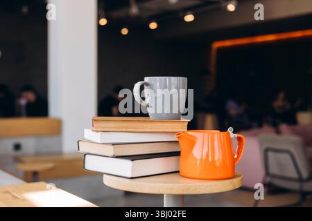 Ein gemütliches Café mit einem Stapel Bücher auf einem Holztisch, begleitet von einem grauen Becher und einer leuchtenden orangefarbenen Teekanne. Der unscharfe Hintergrund fügt ein W hinzu Stockfoto