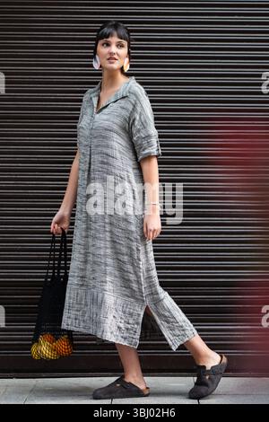 Eine modische Frau spaziert selbstbewusst in der Stadt und trägt dabei eine schwarze Tasche mit Orangen und Bananen. Sie trägt ein stilvolles graues Kleid und schwarze s Stockfoto
