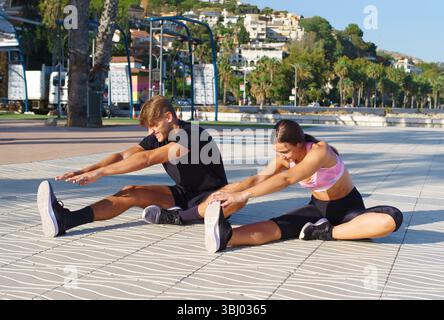 Eine Frau und ein Mann nehmen an Dehnübungen an einer sonnigen Strandpromenade Teil, umgeben von Palmen und einem malerischen Blick auf das Meer und die fernen Hügel Stockfoto