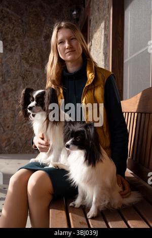 Eine Frau mit gelber Weste sitzt auf einer Holzbank, begleitet von zwei reinrassigen Papillonhunden. Die Hunde zeigen ihre ausgeprägten großen Ohren und s Stockfoto