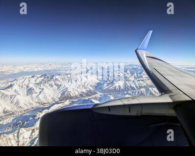 Aus der Vogelperspektive auf schneebedeckte Berge durch ein Flugzeugfenster und ein atemberaubendes Panorama unter einem klaren, hellblauen Himmel. Der Flugzeugflügel Stockfoto