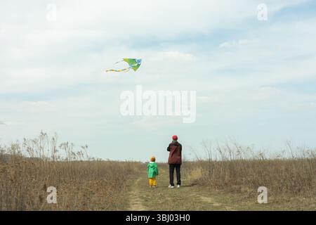 Ein Vater und sein kleiner Sohn teilen einen fröhlichen Moment, in dem sie einen farbenfrohen Drachen auf einem weiten, offenen Feld fliegen. Die Szene fängt das Wesen der Outdoor-Familie ein Stockfoto