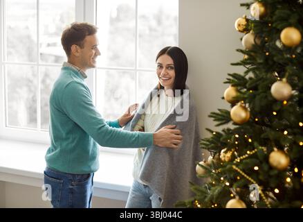 Happy young couple standing by large window and Christmas tree, enjoying festive atmosphere at home. Stockfoto
