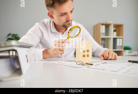 Ein junger Geschäftsmann, der durch eine Lupe auf ein Modellhaus blickt, das am Tisch sitzt Stockfoto