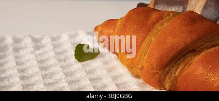 Goldenes Croissant auf einem weißen strukturierten Küchentuch mit grünem Minzblatt. Sanftes Morgenlicht unterstreicht die gemütliche und köstliche Frühstücksumgebung. Stockfoto