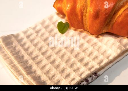 Goldenes Croissant auf einem weißen strukturierten Küchentuch mit grünem Minzblatt. Sanftes Morgenlicht unterstreicht die gemütliche und köstliche Frühstücksumgebung. Stockfoto