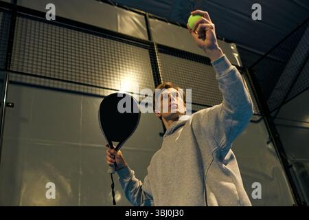 Ein junger, fokussierter Athlet hebt einen Ball auf und ist bereit, auf dem Padel-Platz drinnen zu dienen. Stockfoto