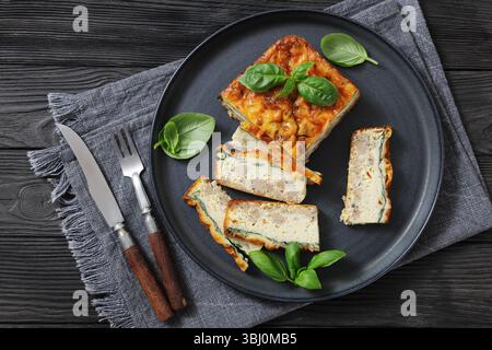 Hausgemachter Wurst-Eierauflauf auf Keramikplatte, garniert mit Basilikum, mit Gabel und Messer auf schwarzem Tisch, echtes Bild von oben Stockfoto