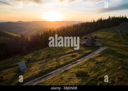 Luftaufnahme eines ruhigen Sonnenuntergangs über hügeliger Landschaft in der Ukraine. Rustikale Holzhütte und unbefestigter Pfad eingebettet zwischen sanften grünen Hügeln, mit Wald und Bergen, die in warmes, goldenes Licht getaucht sind. Stockfoto