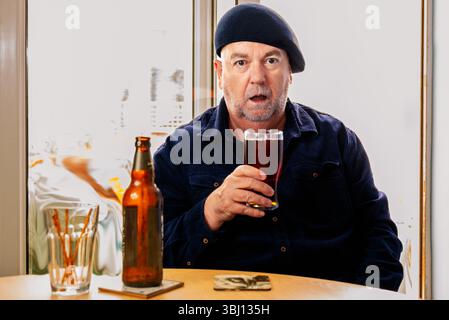 Ein Mann sitzt an einem Tisch mit einer Bierflasche und Brezelstöcken und hält ein Glas dunkles Bier mit einem überraschenden Ausdruck Stockfoto
