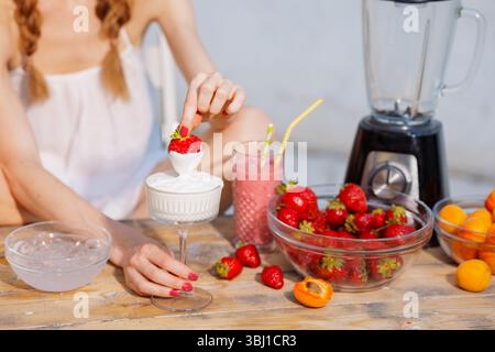 Frau taucht Erdbeere in Creme am Sommertisch mit Smoothie, frischem Obst und Mixer. Gesunder Snack-Moment. Stockfoto