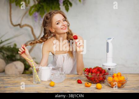 Glückliche Frau mit Zöpfen, die frische Erdbeeren an einem Holztisch mit Obst und Mixer in einer Sommeratmosphäre genießt. Stockfoto