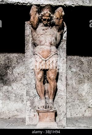 Eine Steinskulptur eines muskulösen Mannes, möglicherweise Atlas, ist dargestellt, der eine Struktur hochhält. Die Skulptur steht an einer strukturierten Wand, mit der des Mannes Stockfoto