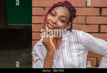 Fröhliche Afroamerikanerin mit roter geflochtener Frisur, die das Kinn nachdenklich berührt. Lifestyle-Porträt im Freien in gestreiftem Hemd an der Ziegelwand Stockfoto