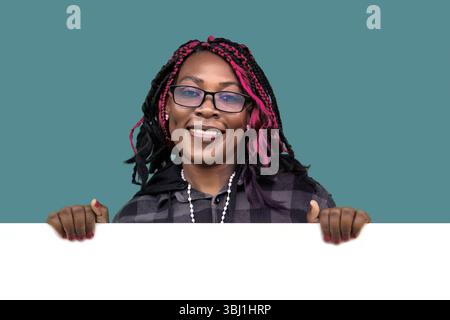 Fröhliche Afroamerikanerin mit Brille und bunten Haaren, die ein weißes Schild hält. Perfekt für Werbung, Werbeaktionen, Ankündigungen und Marketing Stockfoto