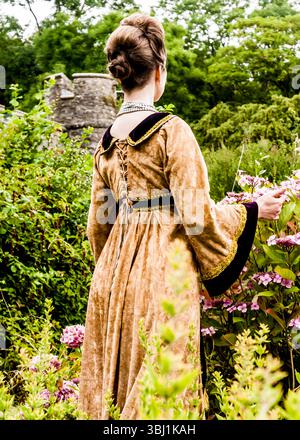 Eine Frau in einem historischen Kostüm steht in einem Garten und berührt die Blumen. Stockfoto