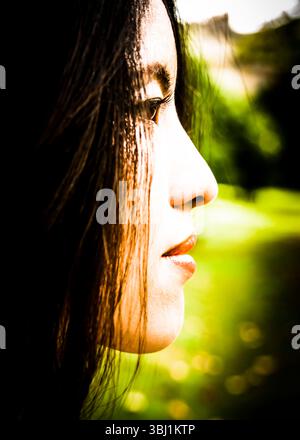 Ein Seitenprofil des Gesichts einer Frau, deren Haare teilweise ihr Gesicht verdecken. Der Hintergrund ist unscharf, mit Grün- und Gelbtönen. Stockfoto