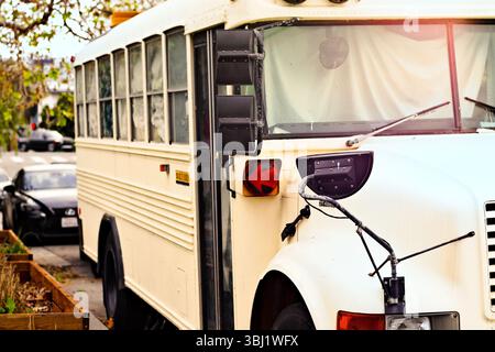 Ein abgenutzter weißer Bus parkt entlang einer ruhigen Nachbarschaftsstraße. Sein Retro-Design und das sanfte warme Licht sorgen für einen nostalgischen urbanen Moment voller sim Stockfoto
