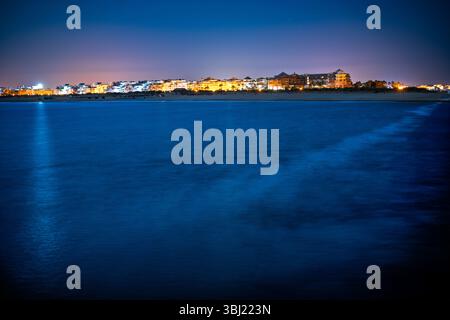 Der ruhige Strand Isla Canela bei Nacht mit sanften Wellen und leuchtenden Gebäuden schafft eine ruhige Atmosphäre. Stockfoto