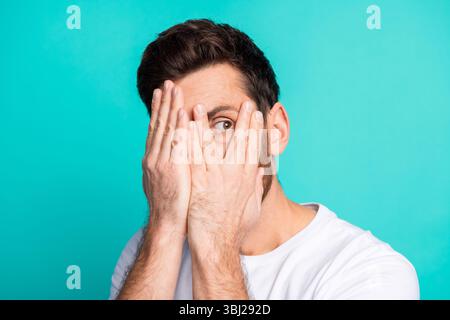 Junger Mann in weißem Hemd, der das Gesicht mit Händen vor blauem Hintergrund bedeckt und Überraschung und Neugier vermittelt Stockfoto