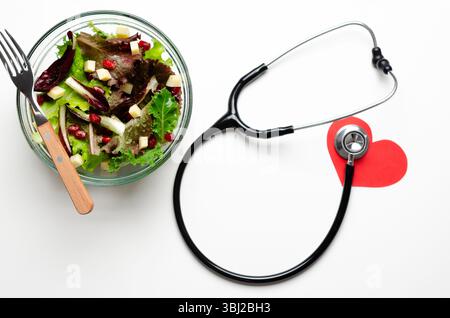 Ein Stethoskop, ein symbolisches rotes Papierherz und ein Salat in einer Glasschale, auf weißem Hintergrund. Healthy Life Concept Draufsicht. Stockfoto