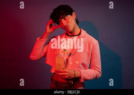 Junge Person, die einen bunten Cocktail hält, eine pinkfarbene Jacke und ein Regenbogenarmband trägt, beleuchtet von roten und blauen Lichtern Stockfoto