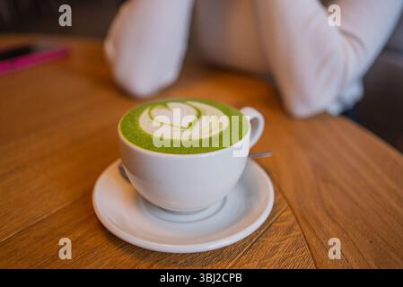 Eine Tasse Matcha Latte mit einem herzförmigen Design aus cremigem Schaum, die auf einem hölzernen Kaffeetisch liegt, schafft eine warme und einladende Atmosphäre, die perfekt zum Entspannen ist Stockfoto