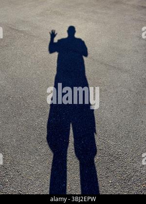 Foto eines großen schwarzen Schattens auf dem Boden eines Asphaltparkplatzes einer stehenden Person aufgrund eines hellen Sonnenlichts, das von hinten strahlt. Stockfoto