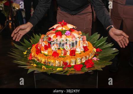 Mehrschichtiger Sushi-Kuchen mit verschiedenen Rollen, belegt mit Tigergarnelen und leuchtenden Blumen auf Bananenblättern. Ein atemberaubendes kulinarisches Herzstück für Hochzeiten Stockfoto