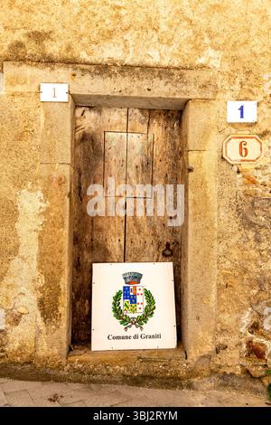 Graniti, Sizilien - Italien - 05-05-2025: Alte Holztür mit verwittertem Rahmen, mit einem weißen Metallschild mit dem bunten Wappen und dem Labyrinth Stockfoto