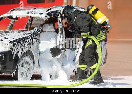 Feuerwehrmann mit Sauerstofftank und Schutzhelm mit weißem Löschschaum, um das Feuer zu löschen, das im zerstörten Auto nach dem ausbrach Stockfoto