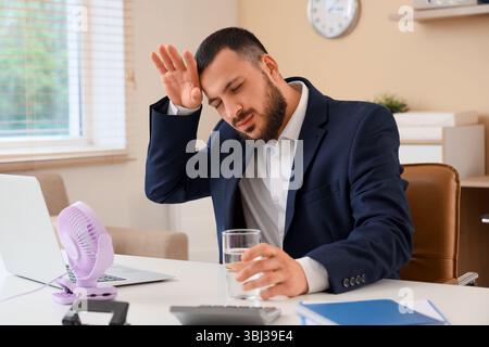 Junger Geschäftsmann mit Glas Wasser und Mini-Ventilator, der am Tisch im Büro unter Hitze leidet Stockfoto