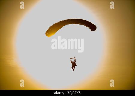 Ein in der Luft befindlicher Soldat der US Army fällt mit Fallschirmen durch den Himmel, während die Sonne während des Abstiegs in einem hellen kreisförmigen Ausbruch direkt hinter ihm leuchtet. Stockfoto