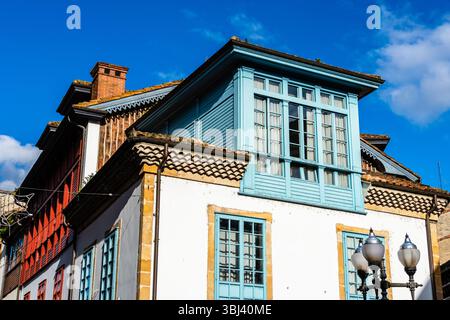 Ein sonnendurchflutetes, blau getrimmtes Erkerfenster ziert ein weißes Gebäude, und die rot getrimmten Fenster auf dem angrenzenden Gebäude sind sichtbar. Villaviciosa, Asturien Stockfoto