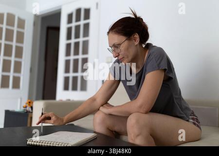 Fokussierte reife Frau mit Brille, die Notizen auf Notizblock schreibt, während sie zu Hause auf dem Sofa sitzt Stockfoto