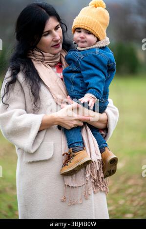 Eine liebevolle Mutter, die Schal und Mantel trägt, hält ihr Baby zärtlich draußen. Das Kind trägt einen Hut und eine Jeansjacke. Beide teilen einen Moment der Wärme und Stockfoto