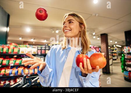 Fröhliche junge Frau in einem blauen Hemd, die Äpfel jongliert, während sie im Supermarkt einkaufen. Umgeben von frischem Obst und einem fröhlichen Lächeln. Konzept von f Stockfoto