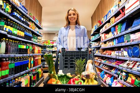 Fröhliche Frau geht einen Supermarktgang mit einem vollen Einkaufswagen entlang, umgeben von farbenfrohen Regalen mit Getränken und Snacks. Das Konzept des Lebensmittelgeschäfts Stockfoto