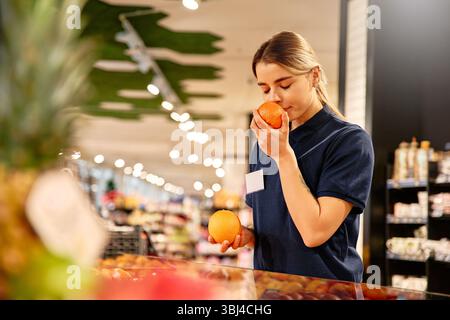 Weibliche Supermarktangestellte riecht orange, während sie einen anderen in der Hand hält. Duftendes Zitrusaroma, Frischekontrolle und lebendige Ladenbeleuchtung betonen Stockfoto
