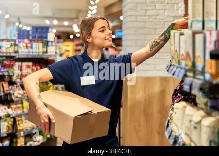Weibliche Angestellte in Marinestraße, die während ihrer Arbeit in einem Lebensmittelgeschäft einen Karton und Lebensmittel hält. Heller, organisierter Gang. Konzept des Einzelhandels Stockfoto