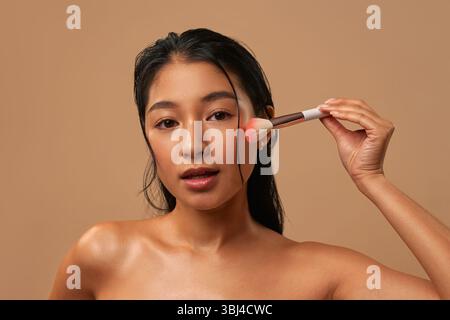 Asian woman with clean healthy glowing skin using makeup brush isolated on beige background. Beauty concept, makeup. Stockfoto